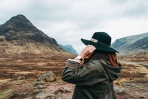 Mujer usando y sosteniendo sombrero de pie contra pintorescas montañas de Escocia - foto de stock