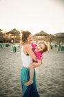 Vista lateral da mulher rindo carregando alegre filho brincalhão a mãos enquanto em pé na praia ao pôr do sol — Fotografia de Stock