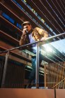 Uomo positivo in abito elegante parlando sul telefono cellulare mentre in piedi sul moderno balcone di vetro di edificio contemporaneo nella giornata di sole — Foto stock