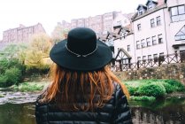 Rückansicht einer Frau mit Hut, die die Landschaft alter gemauerter Gebäude betrachtet, mit seichtem Fluss, der zwischen grünen Büschen fließt, Schottland — Stockfoto