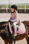 Menina pequena alegre no vestido e jockey feno sentado no cavalo enquanto aprende a montar na pista de corridas — Fotografia de Stock