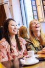 Jóvenes mujeres multirraciales sonriendo y hablando entre sí mientras se sientan en la mesa en el acogedor café - foto de stock
