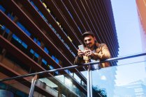Hombre positivo en traje elegante usando el teléfono móvil mientras está de pie en el balcón de cristal moderno del edificio contemporáneo en el día soleado - foto de stock