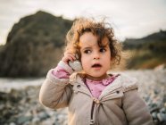 Portrait of cute female kid listening to conch with rapt attention while resting on stony seaside — Stock Photo