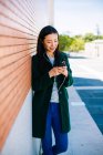 Jeune femme asiatique à l'écoute de la musique et la navigation smartphone tout en s'appuyant sur le mur de briques sur la rue de la ville — Photo de stock