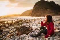 Vista lateral da criança feminina pensativa sentada na praia de pedra e admirando o pôr do sol no fundo da calma beira-mar — Fotografia de Stock