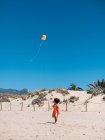 Rückansicht eines kleinen weiblichen Kindes, das am Meer mit fliegendem Drachen vor blauem Himmel rennt — Stockfoto