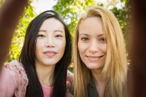 Mulheres jovens multiétnicas sorrindo e olhando para a câmera ao tirar selfie contra árvores verdes no parque — Fotografia de Stock