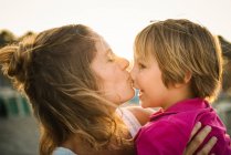 Side view of laughing woman carrying cheerful playful son on hands while kissing his nose on beach in sunset — Stock Photo