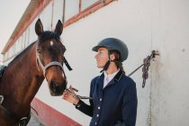 Seitenansicht einer jungen Teenagerin mit Jockeyhelm und Jacke, die ein Pferd streichelt, das zusammen im Freien steht — Stockfoto