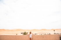 Чудова молода жінка на білому вершечку схилилася на стіні й дивилася на безконечну піщану пустелю (Марокко). — стокове фото