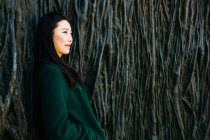 Mujer asiática pensativa en traje de moda mirando hacia otro lado mientras se apoya en la pared con relieve de las raíces de los árboles - foto de stock