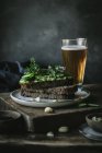 Toasts with green cashew pate, herbs and slices of cucumber with glass of beer on wooden board — Stock Photo