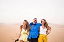 Старший бородатый мужчина смеется и обнимает веселых женщин во время прогулки по песчаной пустыне во время поездки в Марокко — стоковое фото