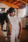 Young woman teenager in white outfit and jockey helmet leading horse out of stall for riding outside — Stock Photo