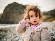 Portrait of cute female kid listening to conch with rapt attention while resting on stony seaside — Stock Photo