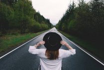 Back view of trendy woman in black hat standing in solitude on remote road with lush green trees, Scotland — Stock Photo
