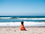 Vista posteriore del bambino femmina in abito arancione seduto sul mare sabbioso e guardando mare e cielo — Foto stock