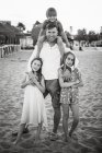 Дорослий чоловік зі смішним хлопчиком на плечах, що стоїть з красивими маленькими дівчатами на пляжі, дивлячись на камеру, чорно-біле фото — стокове фото