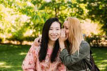 Молода кавказька жінка посміхається і шепоче таємницю до вуха усміхаючись азіатській подрузі, проводячи час у парку разом — стокове фото