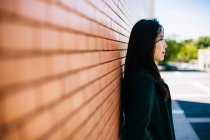 Pensiva donna asiatica in abito alla moda e guardando altrove mentre si appoggia sul muro di mattoni — Foto stock