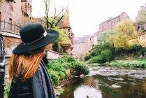 Обратный вид женщины в шляпе, смотрящей на ландшафт старинных каменных зданий с мелкой рекой, текущей среди зеленых кустов, Шотландия — стоковое фото