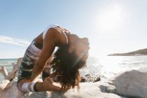 Primer plano de mujer joven atractiva afroamericana que está parada en el puente de la postura del yoga en el fondo del agua calma en día soleado - foto de stock
