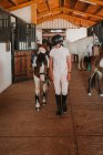 Giovane donna adolescente in abito bianco e casco da fantino che porta il cavallo fuori dalla stalla per andare fuori — Foto stock
