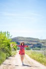 Захоплена молода жінка в модному вбранні розтягує руки і посміхається під час прогулянки по сільській дорозі в сонячний літній день — стокове фото