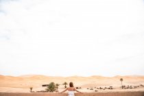 Анонимная молодая женщина в белой верхней части, опирающаяся на стену, смотрящая в сторону бесконечной песчаной пустыни, Марокко — стоковое фото