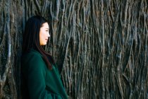 Pensive Asian woman in trendy outfit looking away while leaning on wall with relief of tree roots — Stock Photo