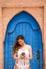 Fröhliche junge Frau trägt weißes Oberteil mit Bikini und fotografiert mit Handy vor blauer orientalischer Tür in Steinmauer, Marokko — Stockfoto