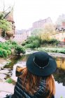 Visão traseira da mulher de chapéu contemplando paisagem de edifícios antigos de alvenaria com rio raso fluindo entre arbustos verdes, Escócia — Fotografia de Stock