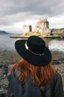 Vista posteriore di donna elegante che indossa un cappello contemplando vecchio castello di pietra sulla costa in montagna, Scozia — Foto stock