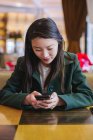 Красивая азиатка просматривает современный смартфон, сидя за столом в уютном кафе — стоковое фото