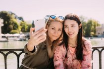 Jóvenes mujeres multirraciales en trajes de moda sonriendo y tomando una selfie con teléfono inteligente mientras se sienta cerca de barandilla de terraplén - foto de stock