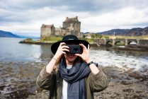Cheerful stylish woman using phone taking photo against old stone castle on coast in mountains, Scotland — Stock Photo