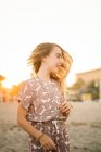Chica sonriente en vestido y accesorios ondeando con el pelo de pie en la playa en la luz del atardecer - foto de stock