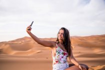 Молода жінка, яка сиділа з телефоном посеред піщаної пустелі (Марокко). — стокове фото