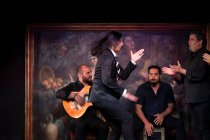 Hombre en traje negro bailando flamenco cerca de músicos masculinos hispanos durante la actuación contra la pintura en el escenario oscuro - foto de stock