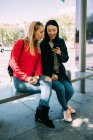 Молодые многорасовые женщины просматривают смартфон, сидя на скамейке автобусной остановки вместе — стоковое фото