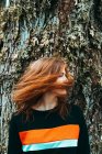 Hermosa mujer joven en sudadera casual ondeando con el pelo de jengibre contra el árbol viejo, Escocia - foto de stock