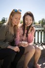 Молоді багаторасові жінки в модних вбраннях посміхаються і переглядають смартфон, сидячи поруч з набережними перилами — стокове фото