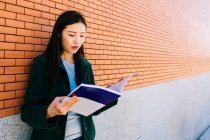 Asiatin liest Lehrbuch, während sie sich auf dem Campus der Universität an eine Ziegelwand lehnt — Stockfoto