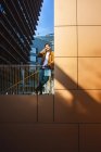 Homem positivo em roupa elegante falando no telefone celular enquanto está em pé na moderna varanda de vidro no edifício contemporâneo no dia ensolarado — Fotografia de Stock