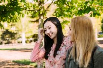 Two multiracial young women in casual outfits talking and looking at each other while sitting on bench in park — Stock Photo
