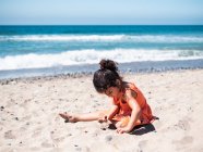 Vista trasera de niña en vestido naranja sentada en la playa de arena y jugando - foto de stock