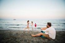 Erwachsene liebende Mann und Frau sitzen am Strand, während ihr Sohn und ihre Töchter zusammen am Meer spielen — Stockfoto