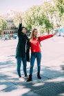 Glückliche multiethnische Damen in stilvollen Outfits, die lächelnd und winkend auf der Straße der Stadt an einem sonnigen Tag zusammen stehen — Stockfoto