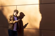 Позитивный человек в стильном наряде с помощью мобильного телефона, опираясь на стену в солнечный день — стоковое фото